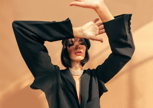 A woman in a black blazer and pearl necklace posing against a tan wall